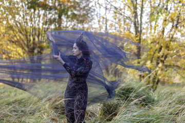 A young woman dressed as a witch and sorceress stands in an autumn forest, wearing a black dress and crown. A beautiful Gothic queen works her magic on Halloween.