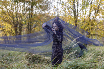 A young woman dressed as a witch and sorceress stands in an autumn forest, wearing a black dress and crown. A beautiful Gothic queen works her magic on Halloween.