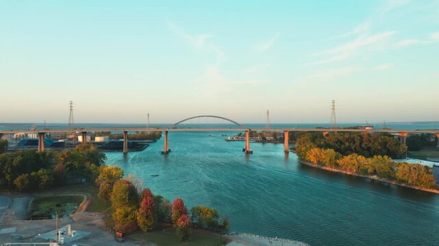 The graceful blue arch of the Leo Frigo Bridge stretches across a broad river framed by autumn trees and industrial docks, glowing gently in the fading dusk light.