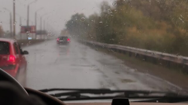 Point of view shot from inside a moving car during a heavy rainstorm. Windshield wipers swipe across the wet glass, revealing blurry traffic and a wet highway asphalt with poor visibility.
