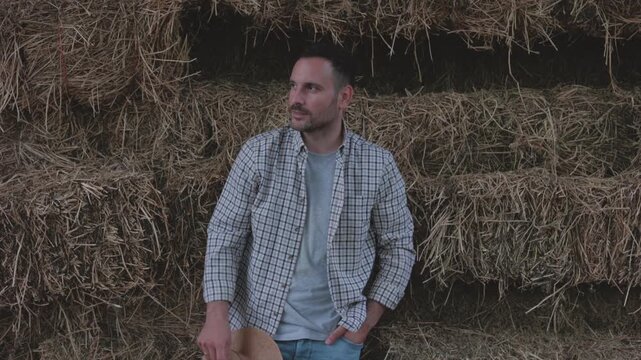 Handsome male farmer in a plaid shirt putting on a straw hat and smiling confidently with arms crossed while leaning against a large stack of hay bales inside a barn or stable after harvest