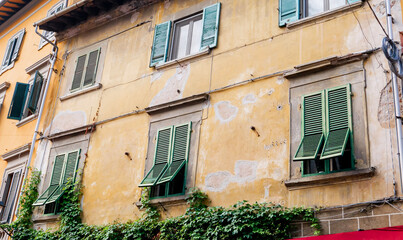 Weathered yellow ochre Italian building facade with green louvered shutters, climbing ivy and peeling plaster in historic Mediterranean town