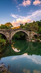 Fototapeta premium An ancient stone bridge arches over calm, reflective water with lush green trees on the sides, under a cloudy blue sky at dusk