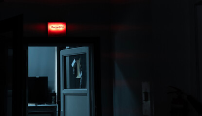 A dark corridor with a slightly open door, above which a red sign reading “RECORDING” is glowing, radio booth