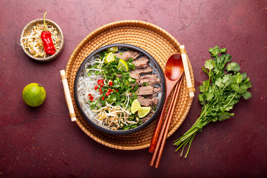Pho Bo Vietnamese soup, bowl of fresh asian soup with rice noodle, beef meat, herbs, soy sprouts and chili on dark red background, top view. Asian cuisine, comfort food.