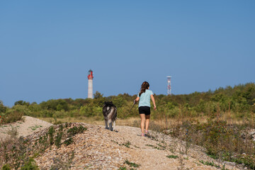Young girl and her Siberian Husky dog hike a scenic coastal trail in Estonia with a lighthouse background.