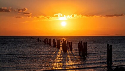 Serene sunset over the ocean with a row of weathered wooden posts casting long reflections on the water and dramatic sun rays breaking through the clouds