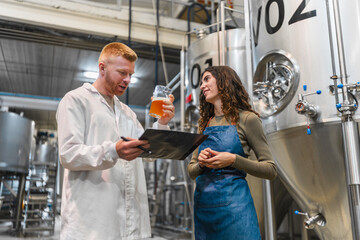 Man and woman in brewery inspecting and tasting beer for quality control, collaborating beside stainless steel fermenting tanks