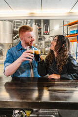 Young couple raising glasses of dark craft beer, smiling at each other during a brewery tour, exploring beer production