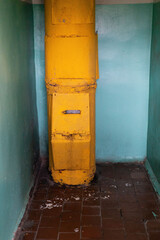 Inside an old building, a yellow garbage chute stands against a wall. The surrounding walls are in a faded blue, and the floor shows signs of wear and dirt