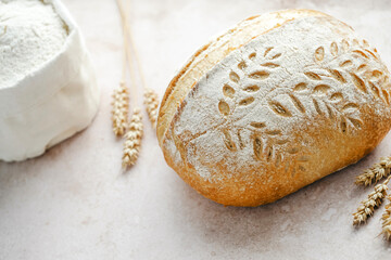 Homemade artisan sourdough loaf is displayed on a light surface, featuring beautiful leaf patterns carved into the crust. Surrounding elements include scattered grains and wheat. 