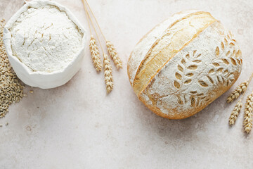 Beautifully baked artisanal bread features a delicate leaf pattern, placed next to a bowl of flour and wheat stalks on a rustic surface, showcasing the art of baking.