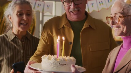 Senior woman taking picture of positive man and surprised birthday lady holding plate with cake, she blowing out candles with cheers from friends during home party