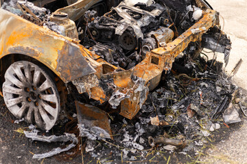 A car wreck lays abandoned by the roadside, heavily burned and charred. The engine compartment is...