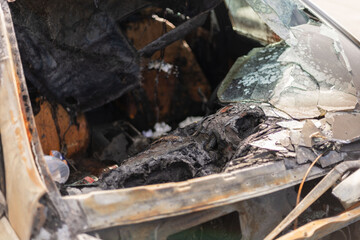 The remains of a burnt car reveal extensive fire damage inside the vehicle's cabin. Charred materials and broken glass create a stark scene in an abandoned location
