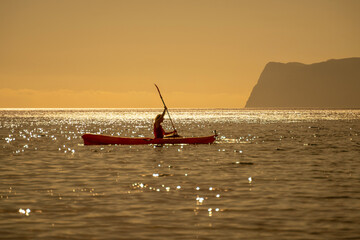 Kayaking, ocean, sunset person paddles kayak on glittering water with distant mountains golden sky...