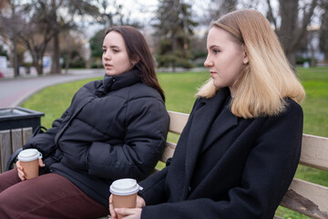 Young women park bench coffee sitting outside together on chilly day drinking takeout beverages