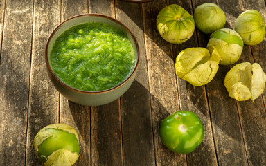 Mexican green salsa (salsa verde cruda) made of fresh tomatillo. Fresh, raw tomatillos in husks and empty husks on the side. Rustic wooden table.