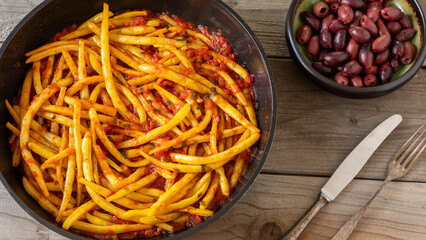 Stewed French beans with tomatoes and olive oil in a pan, a bowl with kalamata olives, a fork, and a knife. Greek cuisine. Rustic wooden table.