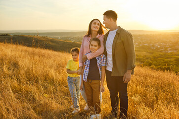 Happy family standing together at sunset, parents cuddle children, husband and wife look at each...