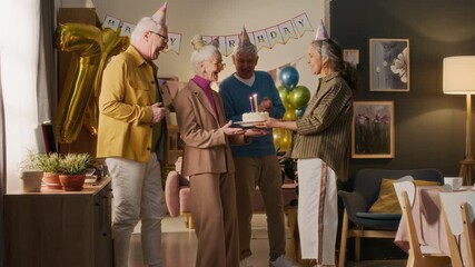 Female friend coming to birthday woman with festive cake, lady blowing out candles while standing between two men, friends clapping hands, all wearing party caps during home celebration