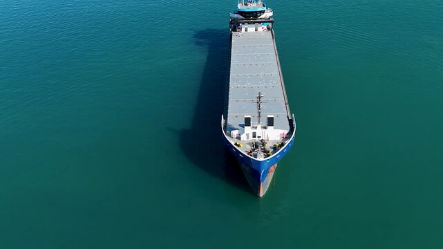 A top down aerial video from a drone looking down at the bow of a blue cargo ship as it moves through the calm water creating a wake