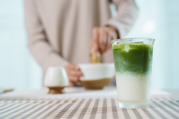 Close up of iced matcha latte on table with Asian woman making tea behind.