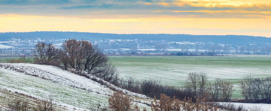 Panoramic rural landscape at sunset in late autumn or early winter with hills covered with first snow, and a green winter crop field against the backdrop an orange-blue sky