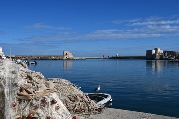A seagull stands on the prow of a boat near a pile of fishing nets. The water and sky are a dark blue. Stone walls surround the harbour, and a red lighthouse is in the distance.