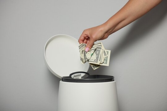 Woman throwing dollar banknotes into trash bin near light wall indoors, closeup