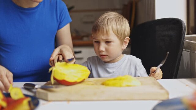 Mother teaching her curious son how to cut mango fruit at home kitchen