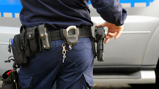 Police officer in a uniform with his hand on holstered pistol, duty belt carrying handcuffs, pepper spray, keys, standing near patrol vehicle during urban response, generic shot, at the ready, Poland