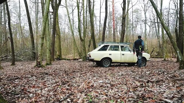 A man and his black dog alongside an old station wagon with a roof rack, parked in a rustic, leafless forest covered in dry leaves. Perfect for nature, travel, or vintage adventure themes.