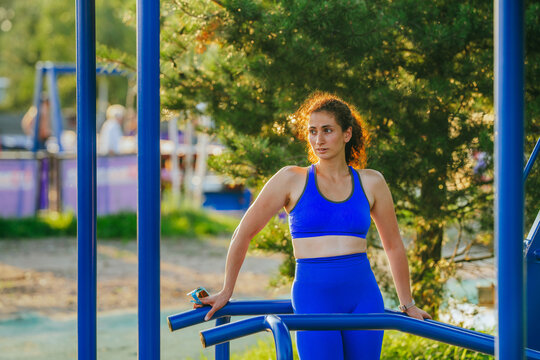 A woman in blue athleisure rests between parallel bars under the afternoon sun, surrounded by greenery and an outdoor gym.