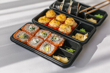 Assorted sushi rolls in black plastic containers with wasabi, pickled ginger and chopsticks on a white table, concept of japanese cuisine, takeaway food and healthy eating