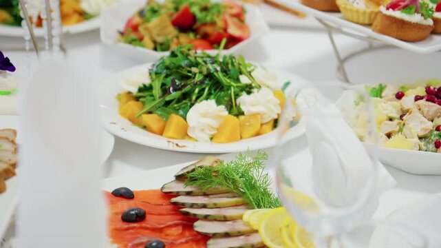 Assorted appetizers on buffet table: salads, sliced salmon and cold cuts, canapes and rolls, white table setting