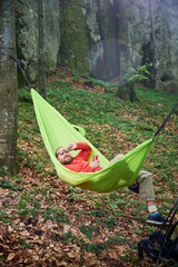 Person relaxing in bright green hammock tied between two trees in lush, green forest near rocks....