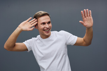 Smiling man in casual white T-shirt facing camera, raising hands in friendly greeting gesture,...