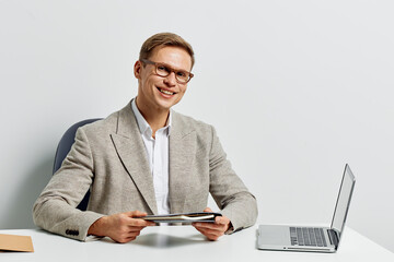 Professional smiling man with glasses in beige blazer sitting at desk with laptop and tablet in modern office environment, looking at camera, happy expression, business casual attire, professional