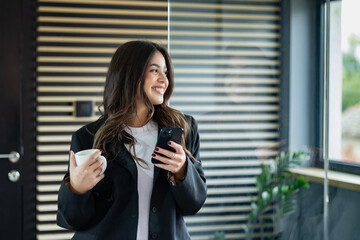 A smiling young businesswoman stands in an office setting, holding a coffee mug while engaged with her smartphone © Panorama