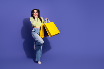 Fototapeta premium Excited young woman in hip casual outfit holding yellow shopping bags, gesturing joy against a vivid purple backdrop