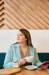 Woman sitting at table holding cup of coffee and smartphone, looking away with slight smile,...