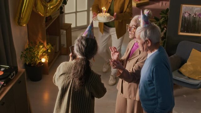 High angle shot of company of senior friends surprising delighted lady with festive cake and clapping to celebrate her 70th birthday, woman taking smartphone photos