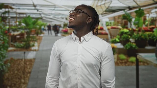 Man smiles and looks up at colorful flowers in a street market, face turned and hands relaxed, wearing white shirt; joy community.
