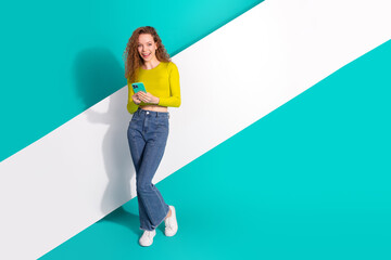 Smiling young woman holding a smartphone, wearing a vibrant yellow top and denim jeans, leaning...