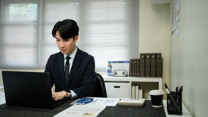 Fototapeta premium Asian business man in suit using laptop and tablet while sitting at her working place. Concentrated at work.