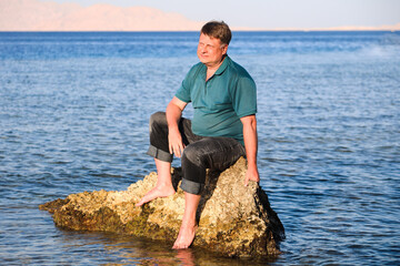 A man in clothes sits on a rock in the sea far from the shore.