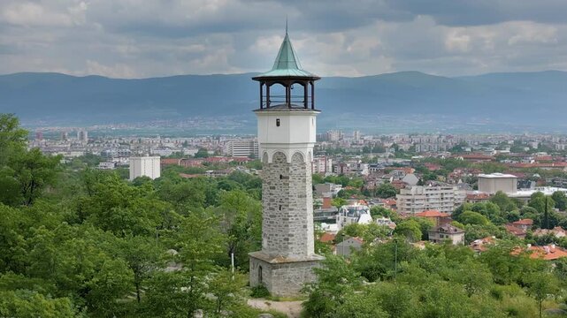 Aerial orbit shot of Plovdiv&rsquo;s iconic clock tower on Sahat Tepe (Danov Hill), surrounded by green trees under a cloudy spring sky, with the city and mountains in the background