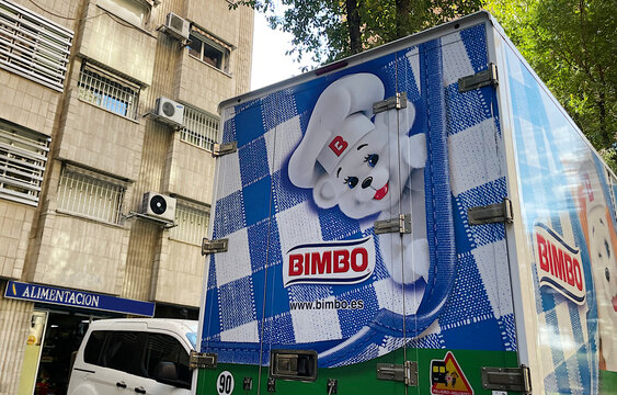 Cami&oacute;n proveedor de reparto de Bimbo con logo de oso con delantal y gorro de panadero en doble fila suministrando pan y boller&iacute;a a comercio de alimentaci&oacute;n en la calle