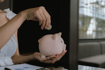 A woman putting coins into a pink piggy bank, symbolizing saving money, financial planning, and...